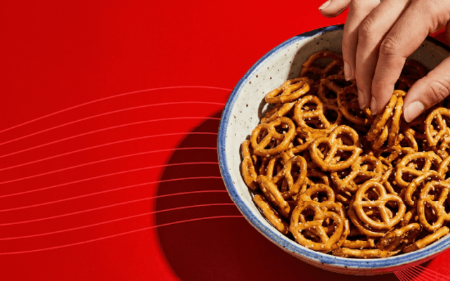 A hand reaching into a blue-rimmed bowl filled with golden mini pretzels against a bold red background.